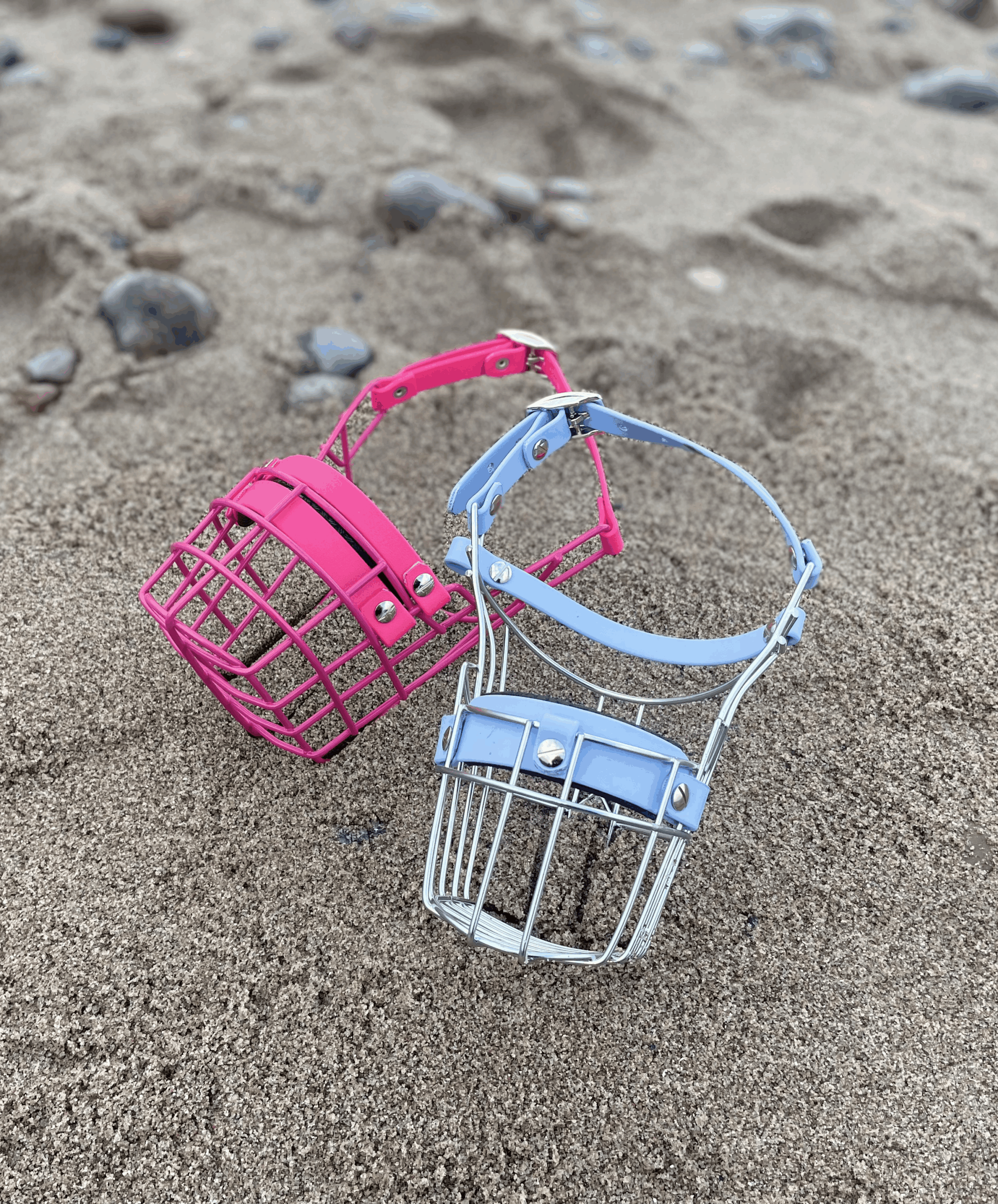 Zwei Maulkörbe im Sand. Einer ist pink, der andere Himmelblau.