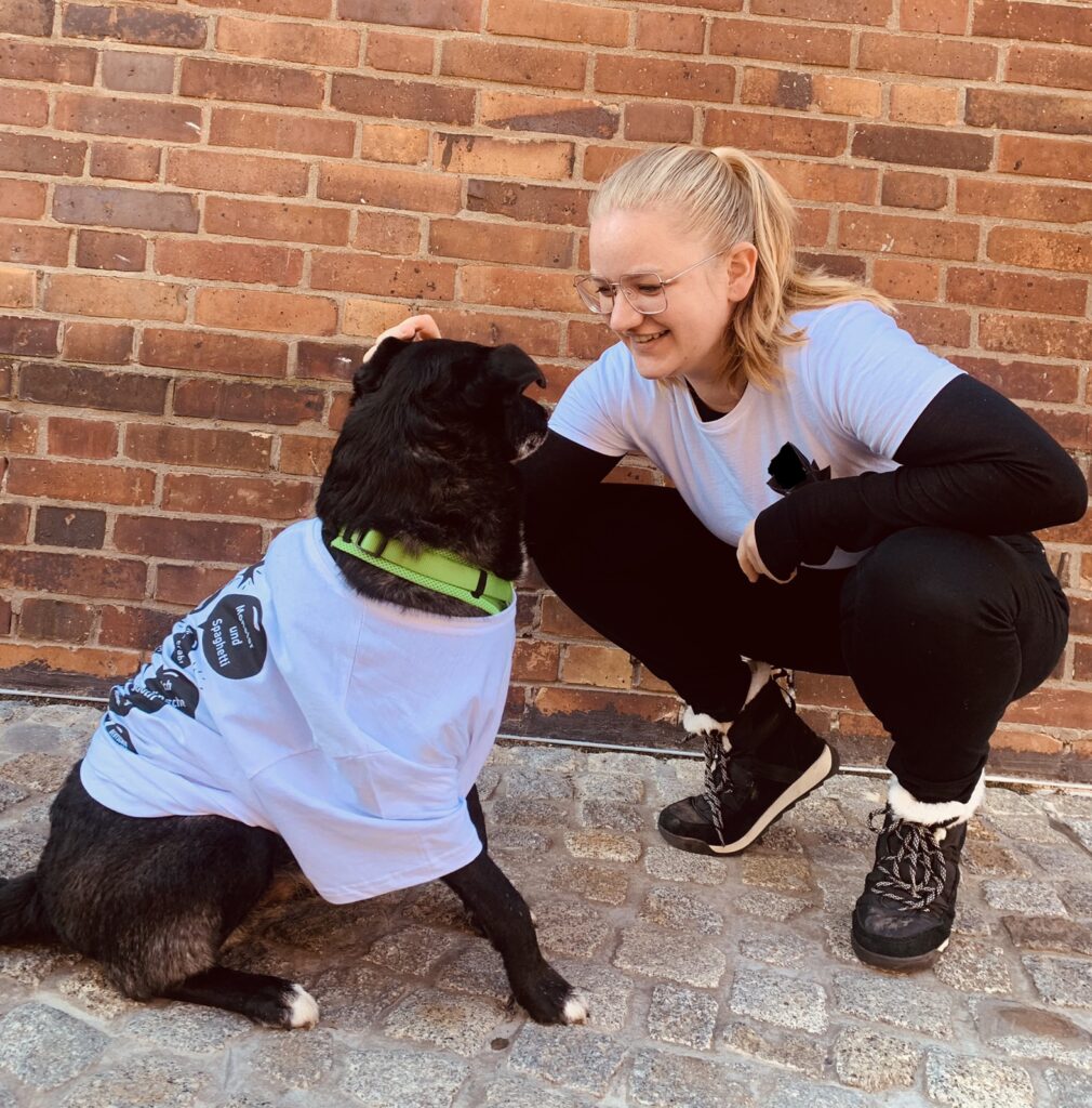 Auf dem Bild sind ein schwarzer Hund und eine blonde Frau zusehen. Die Frau ist in der Hocke. Beide sind einander zugewandt. Sie tragen jeweils ein weißes T-Shirt.